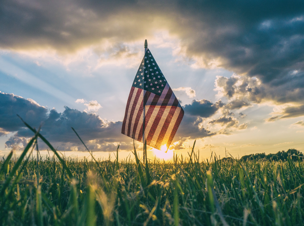 alt="American flag stands in grass field during sunset"